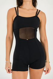 A woman models the Gia Unitard—a fitted black romper with double shoulder straps and sheer mesh midriff panels—accessorized with gold jewelry, against a plain light background.