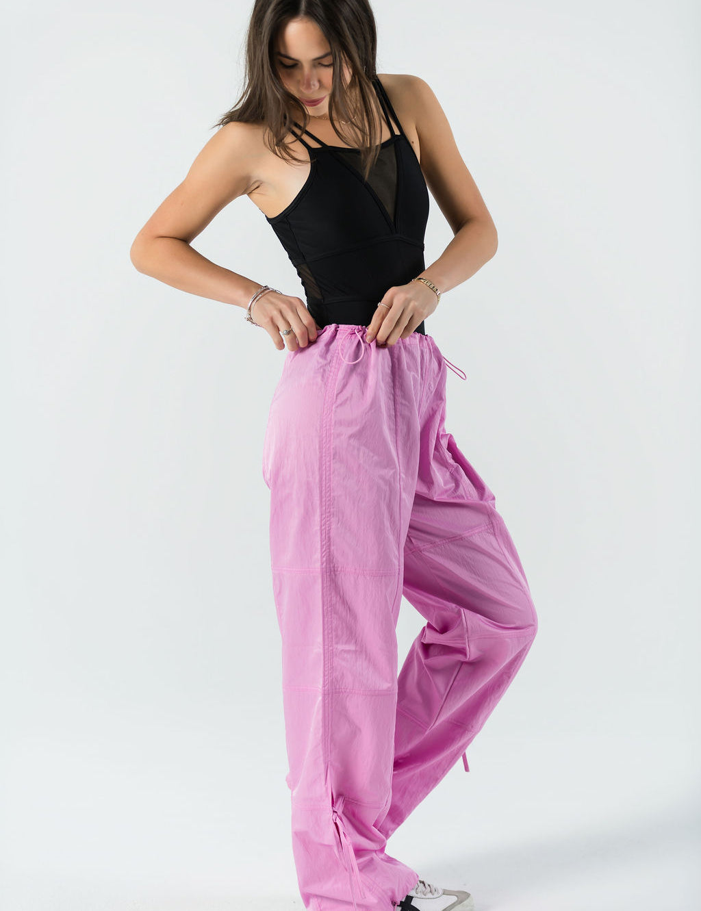 A woman stands against a white background, wearing WOW Parachute Pants in pink with an adjustable waist and a black sleeveless top, looking down as she adjusts the waistband.