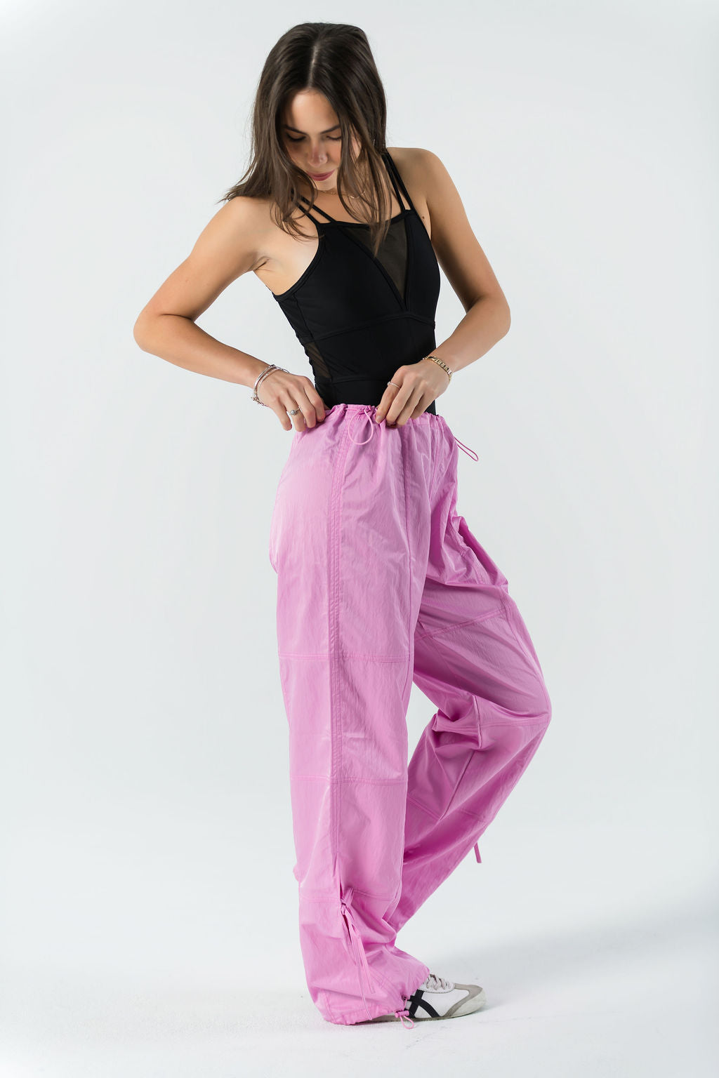 A woman stands against a white background, wearing WOW Parachute Pants in pink with an adjustable waist and a black sleeveless top, looking down as she adjusts the waistband.