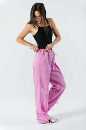 A woman stands against a white background, wearing WOW Parachute Pants in pink with an adjustable waist and a black sleeveless top, looking down as she adjusts the waistband.