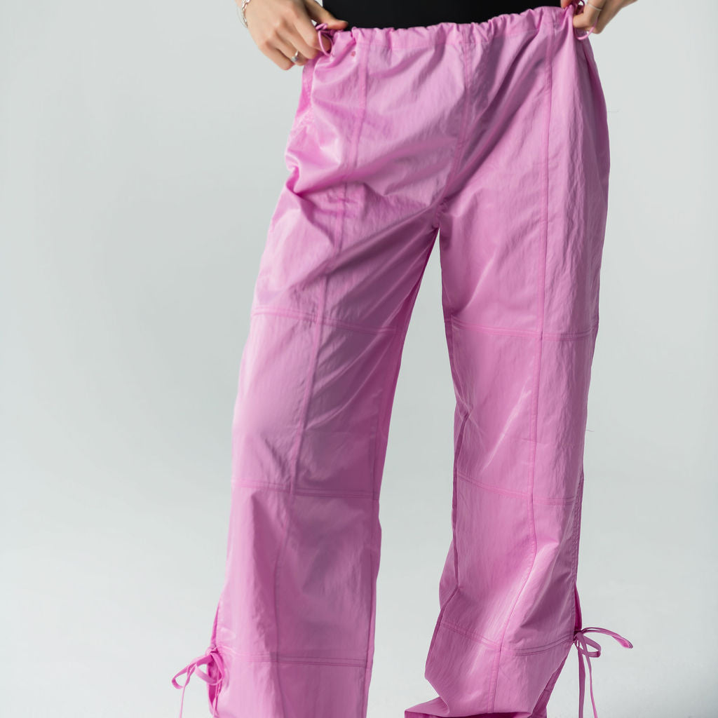 A person wearing a sleeveless black top, white sneakers, and WOW Parachute Pants in pink—with an adjustable waist and ankle drawstrings—stands against a plain light background, holding the waistband of the pants.