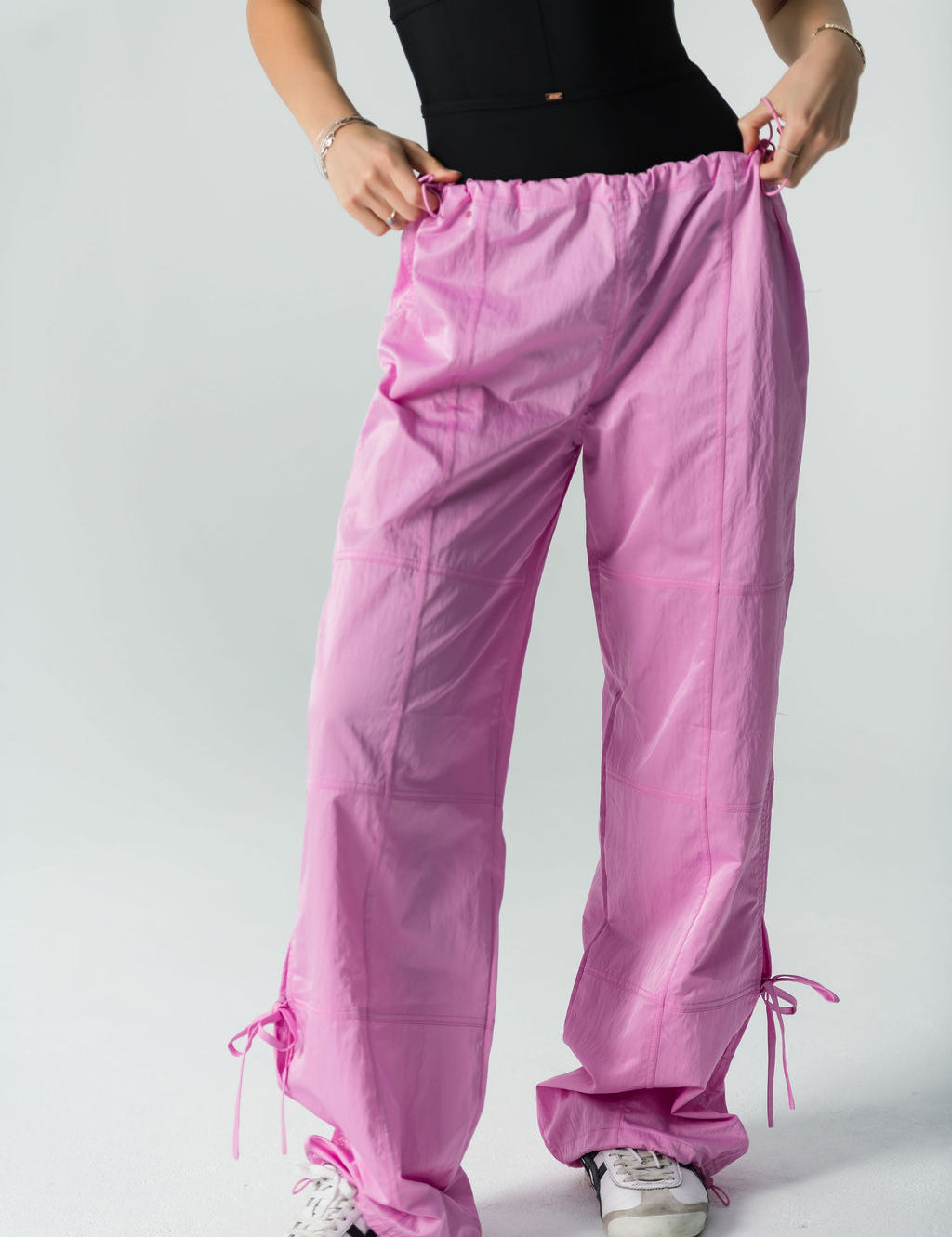 A person wearing a sleeveless black top, white sneakers, and WOW Parachute Pants in pink—with an adjustable waist and ankle drawstrings—stands against a plain light background, holding the waistband of the pants.