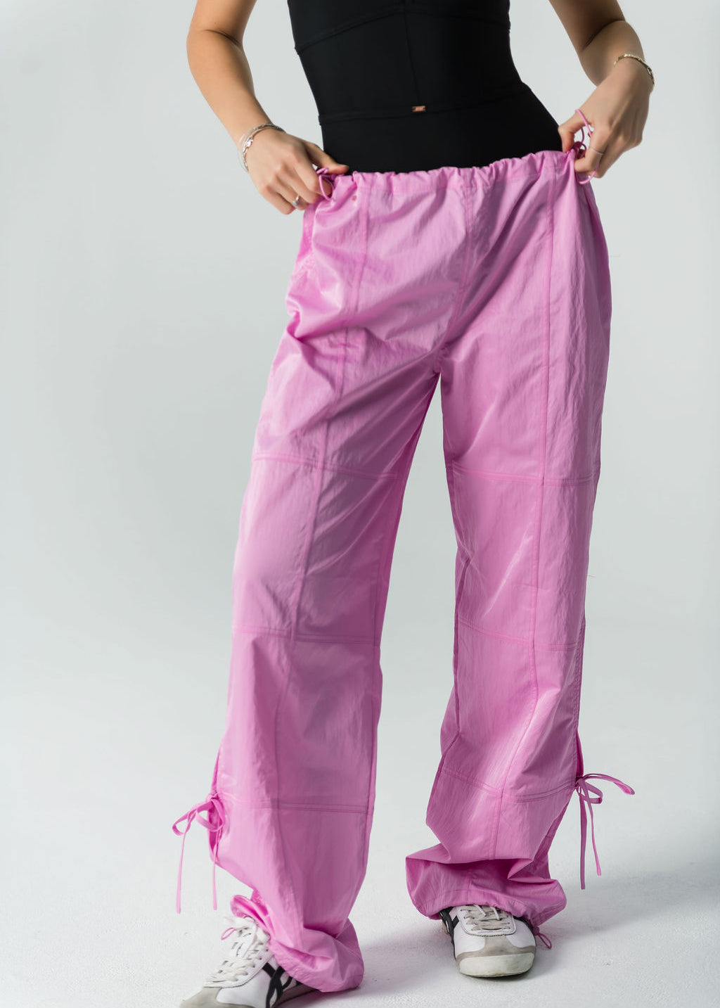 A person wearing a sleeveless black top, white sneakers, and WOW Parachute Pants in pink—with an adjustable waist and ankle drawstrings—stands against a plain light background, holding the waistband of the pants.
