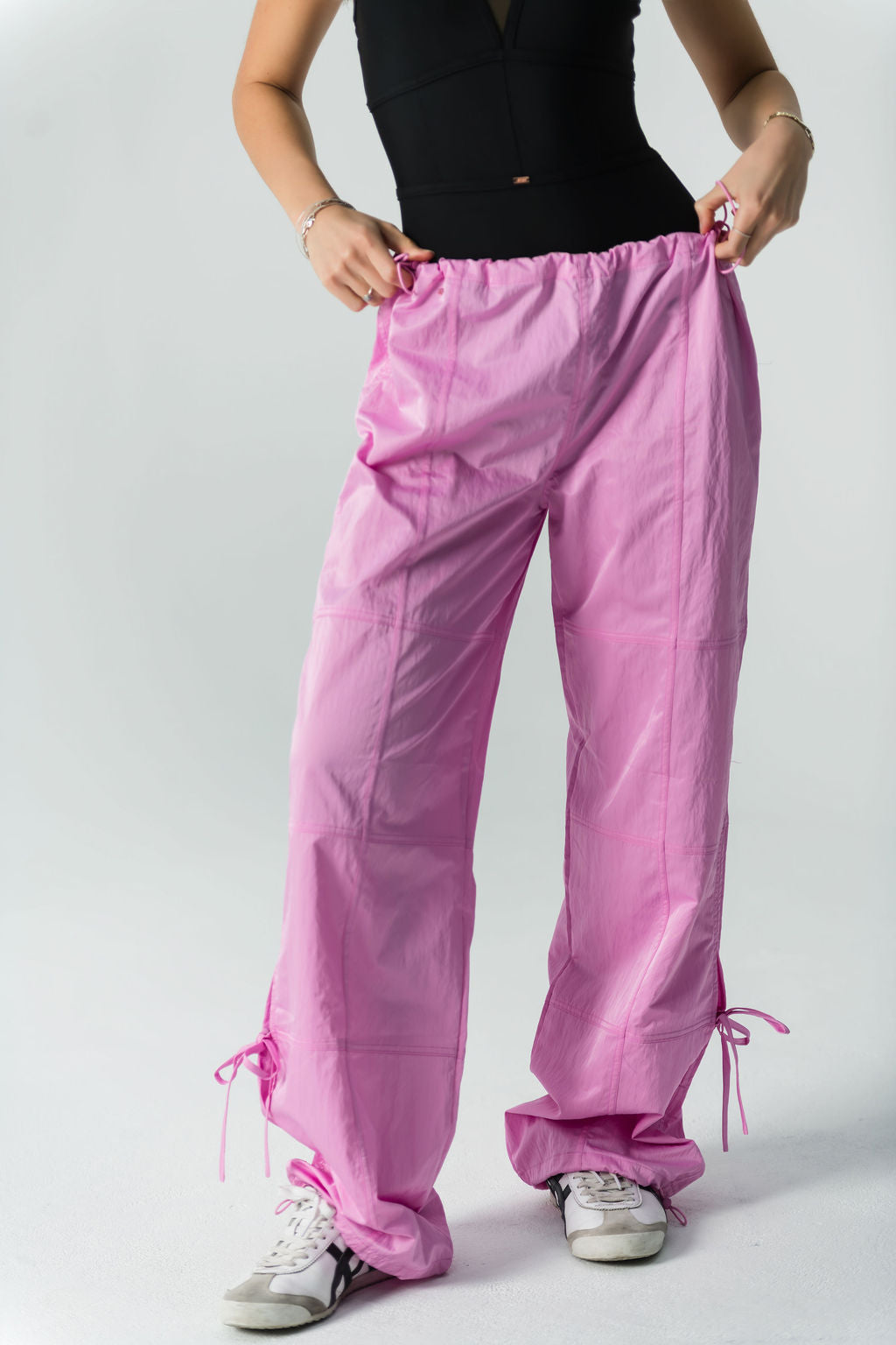 A person wearing a sleeveless black top, white sneakers, and WOW Parachute Pants in pink—with an adjustable waist and ankle drawstrings—stands against a plain light background, holding the waistband of the pants.