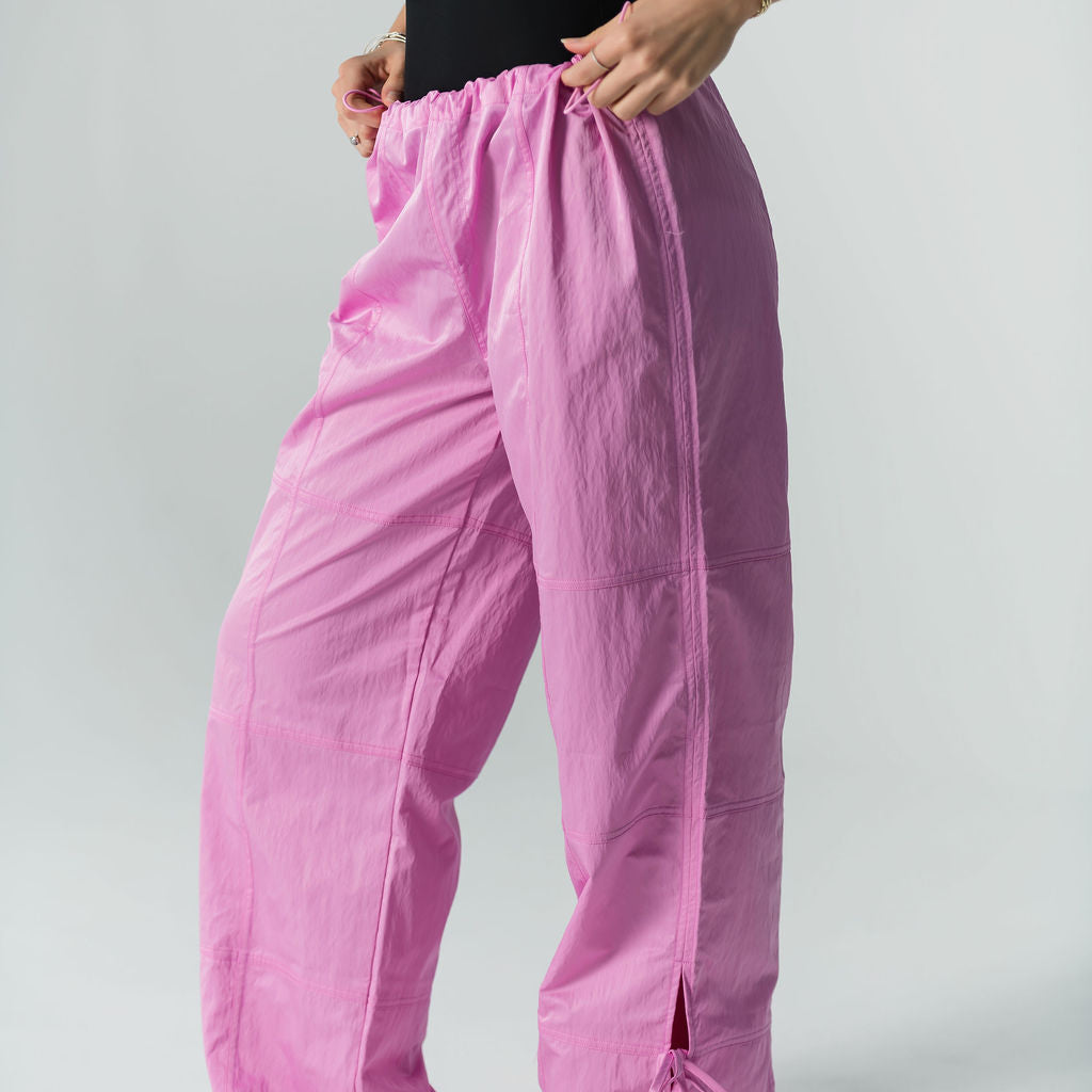 Someone models WOW Parachute Pants in bright pink, featuring an adjustable waist and drawstrings, paired with a black sleeveless top and white sneakers, against a plain light background.