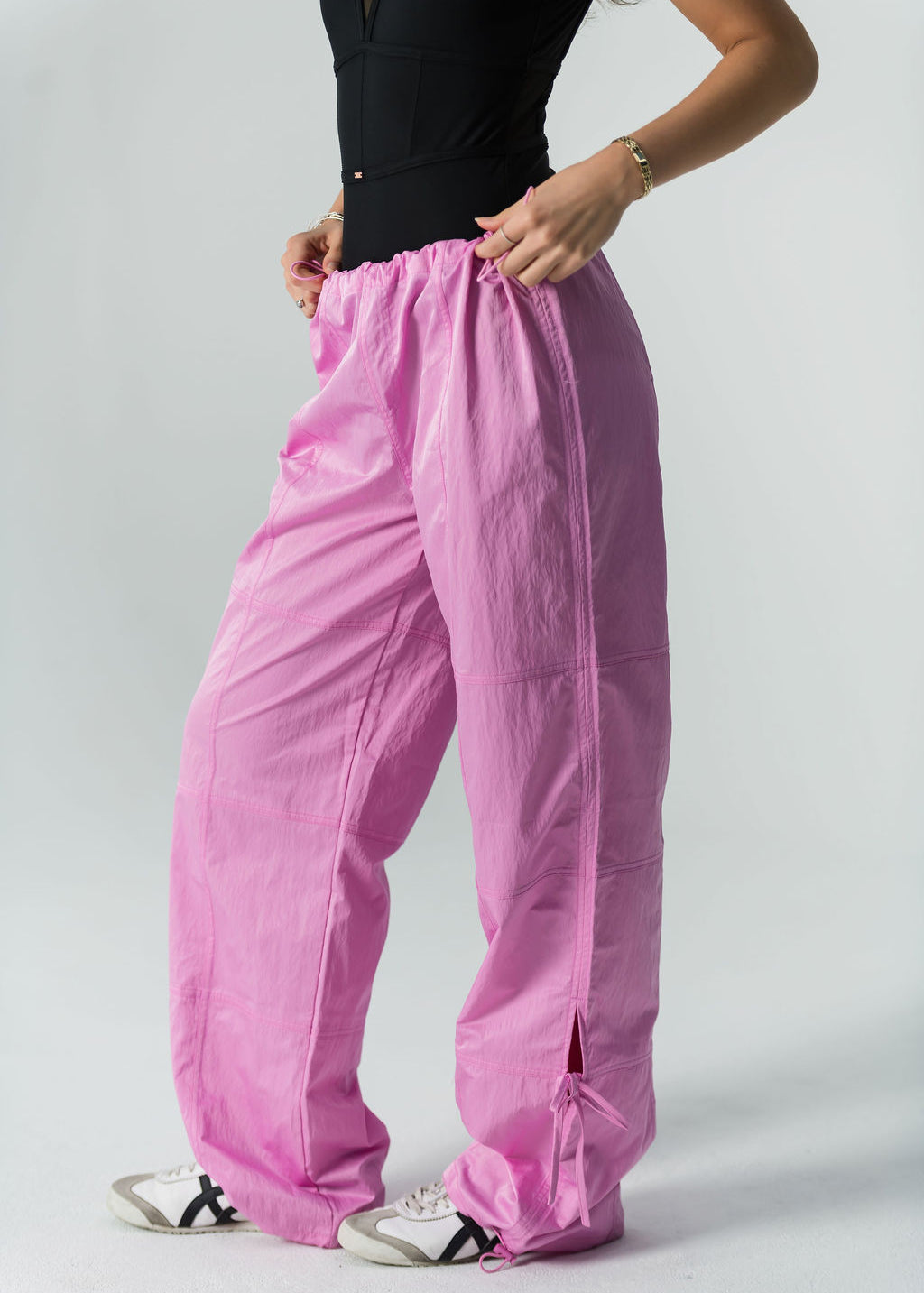 Someone models WOW Parachute Pants in bright pink, featuring an adjustable waist and drawstrings, paired with a black sleeveless top and white sneakers, against a plain light background.