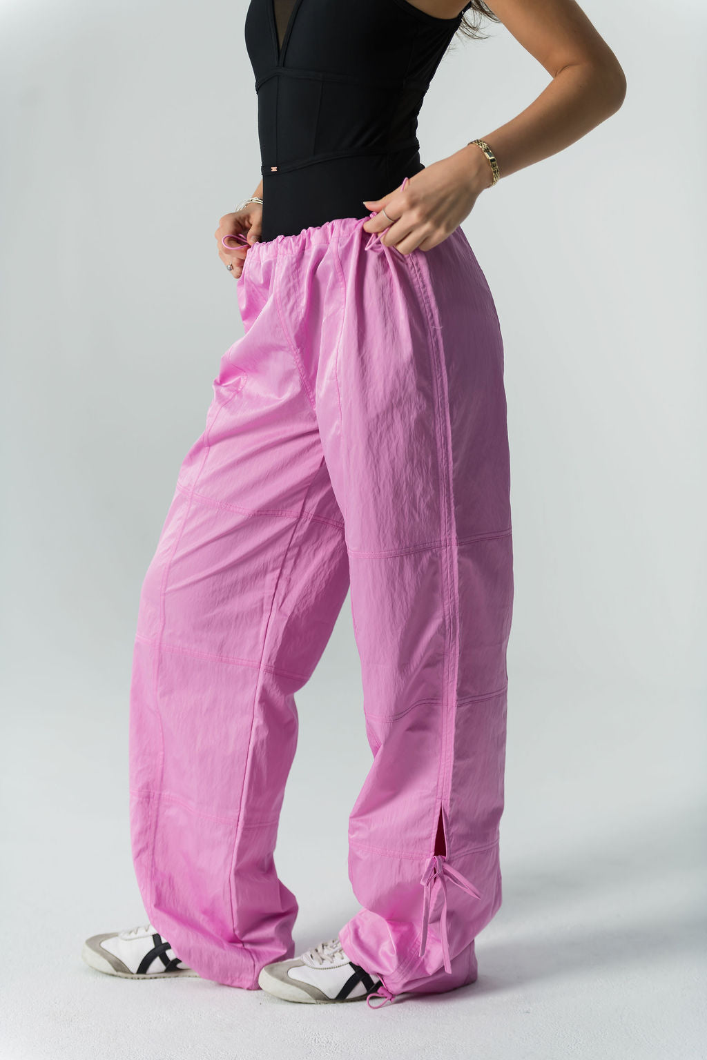 Someone models WOW Parachute Pants in bright pink, featuring an adjustable waist and drawstrings, paired with a black sleeveless top and white sneakers, against a plain light background.