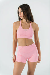A woman stands against a white background, wearing the Bonbon Top in Vivid Pink featuring a mesh scoop neckline and matching high-waisted athletic shorts. She looks to the side with her arms relaxed at her sides.