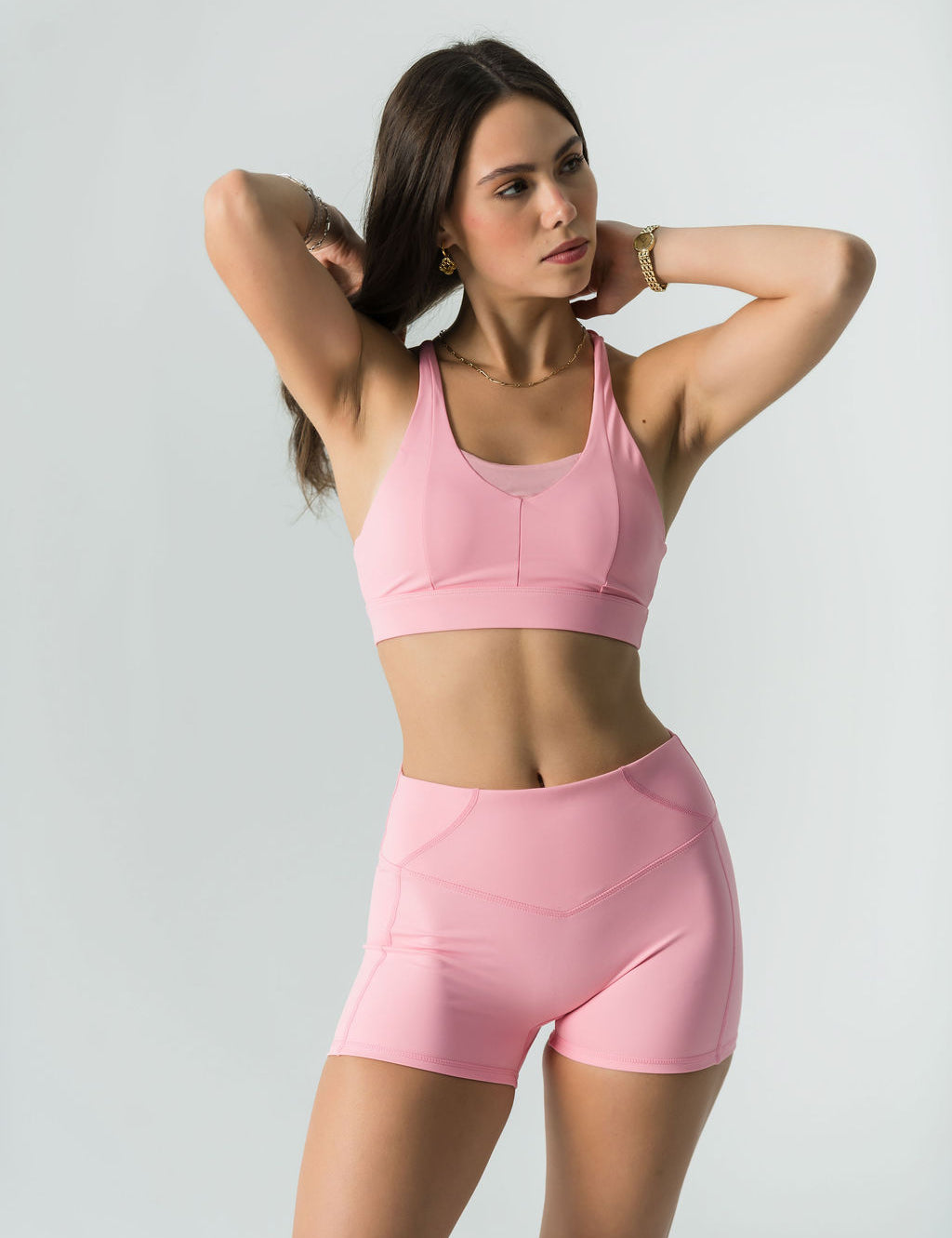 A woman wears the Bonbon Shorts in Vivid Pink, made from soft, stretchy fabric, and poses with her hands behind her head against a plain light background.