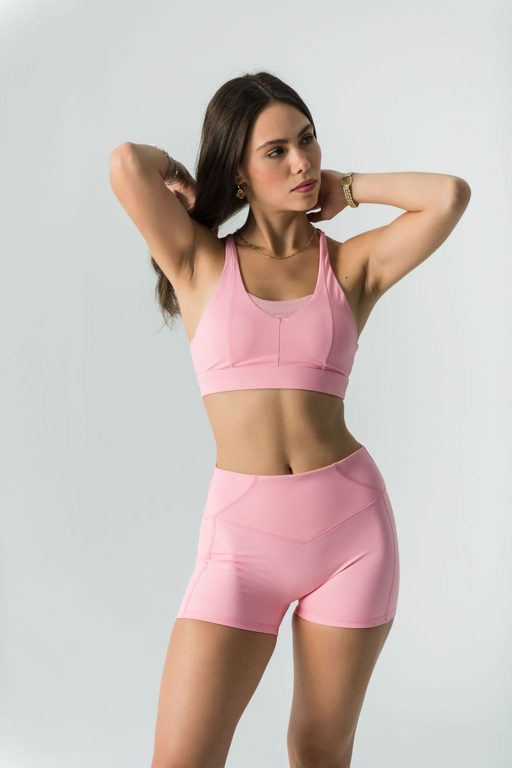A woman wears the Bonbon Shorts in Vivid Pink, made from soft, stretchy fabric, and poses with her hands behind her head against a plain light background.