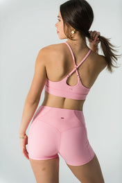 A woman with long brown hair in a ponytail wears the Bonbon Top in Vivid Pink and shorts. She faces away, looking over her shoulder and holding her ponytail to display the adjustable crisscross straps against a plain light background.