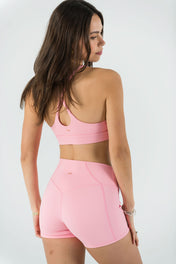 A woman with long brown hair models the Bonbon Shorts in Vivid Pink and a light pink sports bra, made from soft, stretchy fabric, posing with her back to the camera against a plain white backdrop.