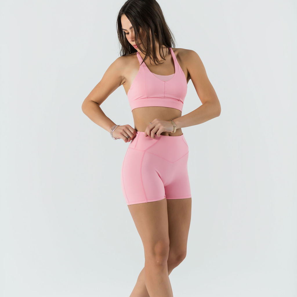 A woman in a light pink sports bra and Bonbon Shorts - Vivid Pink stands against a white background, looking down as she adjusts her seamless shorts. She wears white sneakers and has long, loose hair.