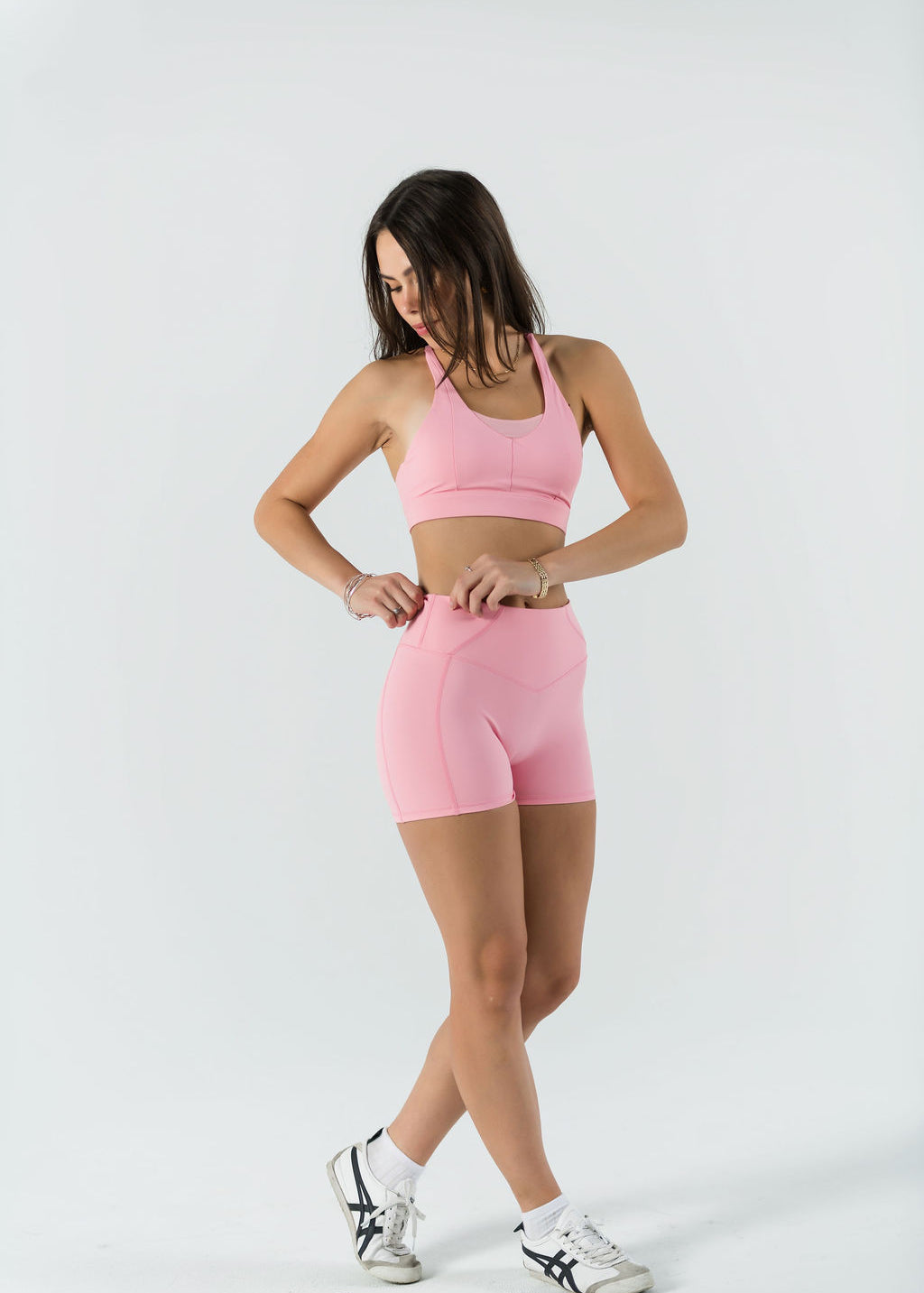 A woman in a light pink sports bra and Bonbon Shorts - Vivid Pink stands against a white background, looking down as she adjusts her seamless shorts. She wears white sneakers and has long, loose hair.