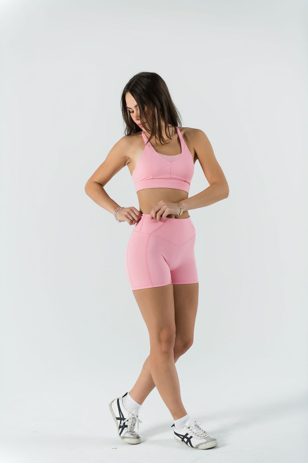A woman in a light pink sports bra and Bonbon Shorts - Vivid Pink stands against a white background, looking down as she adjusts her seamless shorts. She wears white sneakers and has long, loose hair.