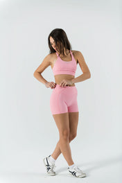 A woman in a light pink sports bra and Bonbon Shorts - Vivid Pink stands against a white background, looking down as she adjusts her seamless shorts. She wears white sneakers and has long, loose hair.