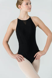 A young woman wears the AVA Leotard in black, featuring a crisscross back, with light tights. She stands hands on hips and gazes to the side against a plain light background.