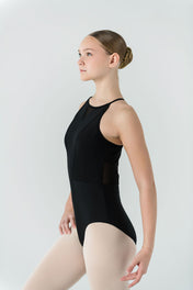 A young ballet dancer with a bun wears the black AVA Leotard featuring a crisscross back and light tights, standing in profile with her arm slightly raised against a plain light background.