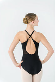 A ballet dancer faces away, wearing the AVA Leotard with a black crisscross open back, light tights, and a neat bun. Her hands rest on her hips against a plain white background.