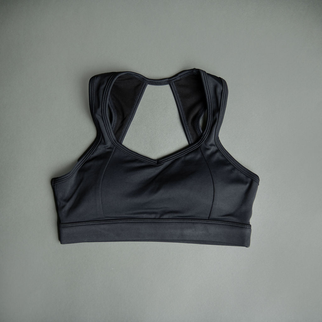 The Allure Top - Black, a racerback sports bra with contrast stitching, is laid flat on a gray surface displaying its front and back straps.
