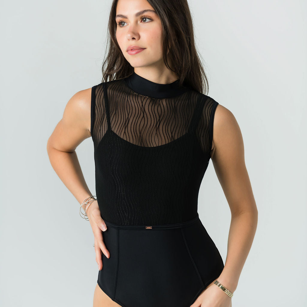A woman poses confidently against a light gray background in Eden's Leo - Bold Edition, a luxury sleeveless black leotard featuring a sheer wavy-patterned mesh top. She stands with one hand on her hip, looking slightly to the side.