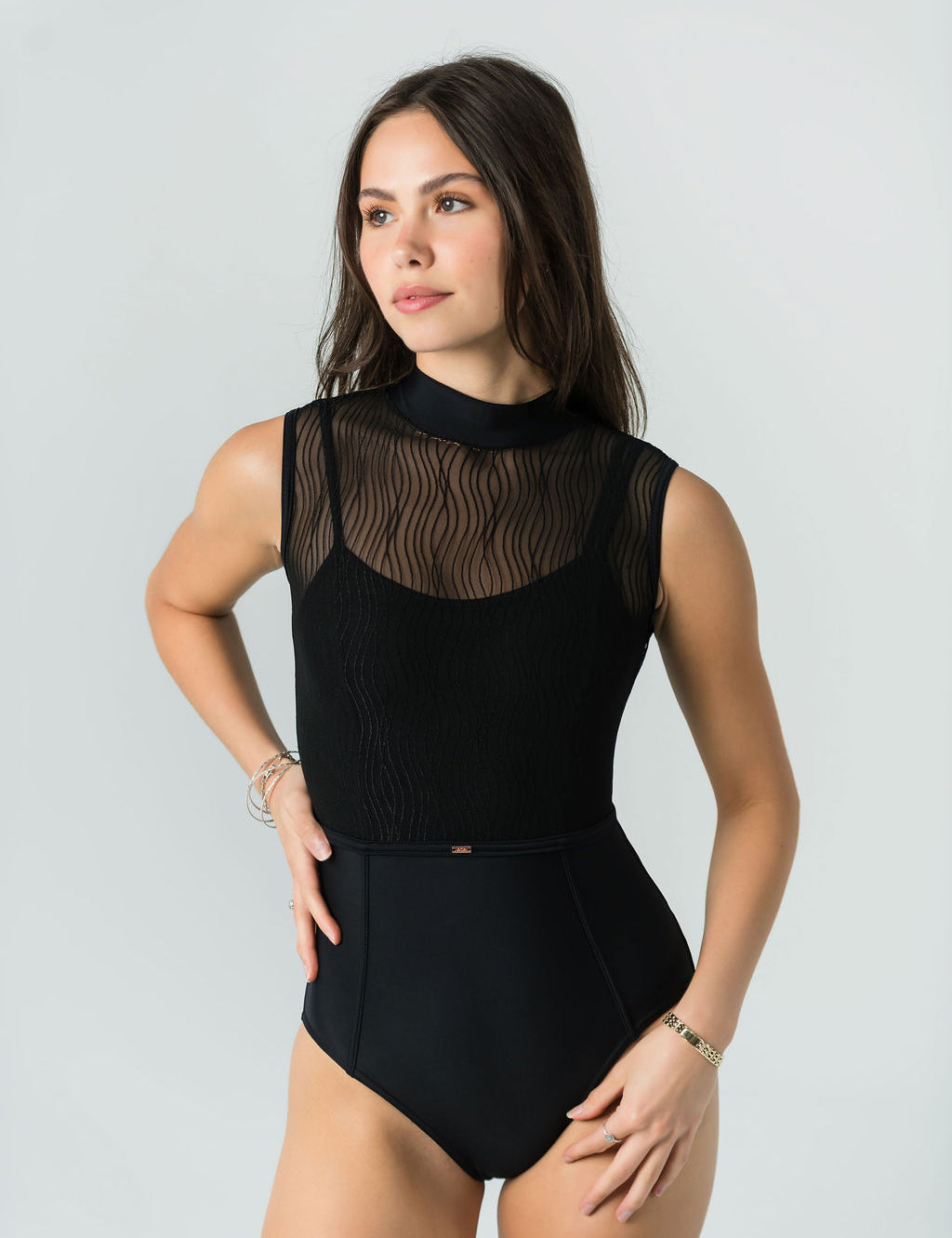 A woman poses confidently against a light gray background in Eden's Leo - Bold Edition, a luxury sleeveless black leotard featuring a sheer wavy-patterned mesh top. She stands with one hand on her hip, looking slightly to the side.