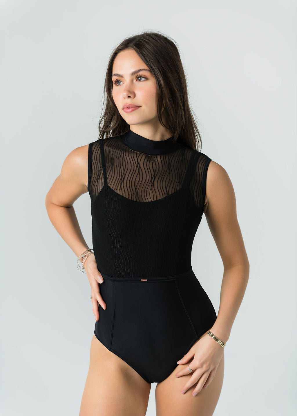 A woman with long brown hair poses against a light gray background in the Eden Leotard - Bold Edition, a black luxury activewear piece featuring a sheer, patterned mesh top. She stands with one hand on her hip, gazing slightly to the side.