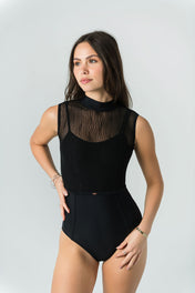 A woman poses confidently against a light gray background in Eden's Leo - Bold Edition, a luxury sleeveless black leotard featuring a sheer wavy-patterned mesh top. She stands with one hand on her hip, looking slightly to the side.