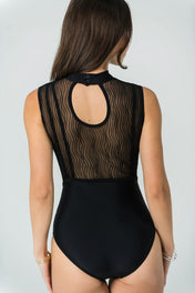 A woman with long brown hair wears Eden's Leo - Bold Edition, a sleeveless black high neck leotard featuring a sheer, wavy mesh back and keyhole cutout. She accessorizes with a bracelet for a chic luxury activewear look.